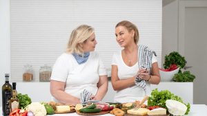 Two women in a kitchen preparing fresh vegetables and cheese with olive oil and spices.